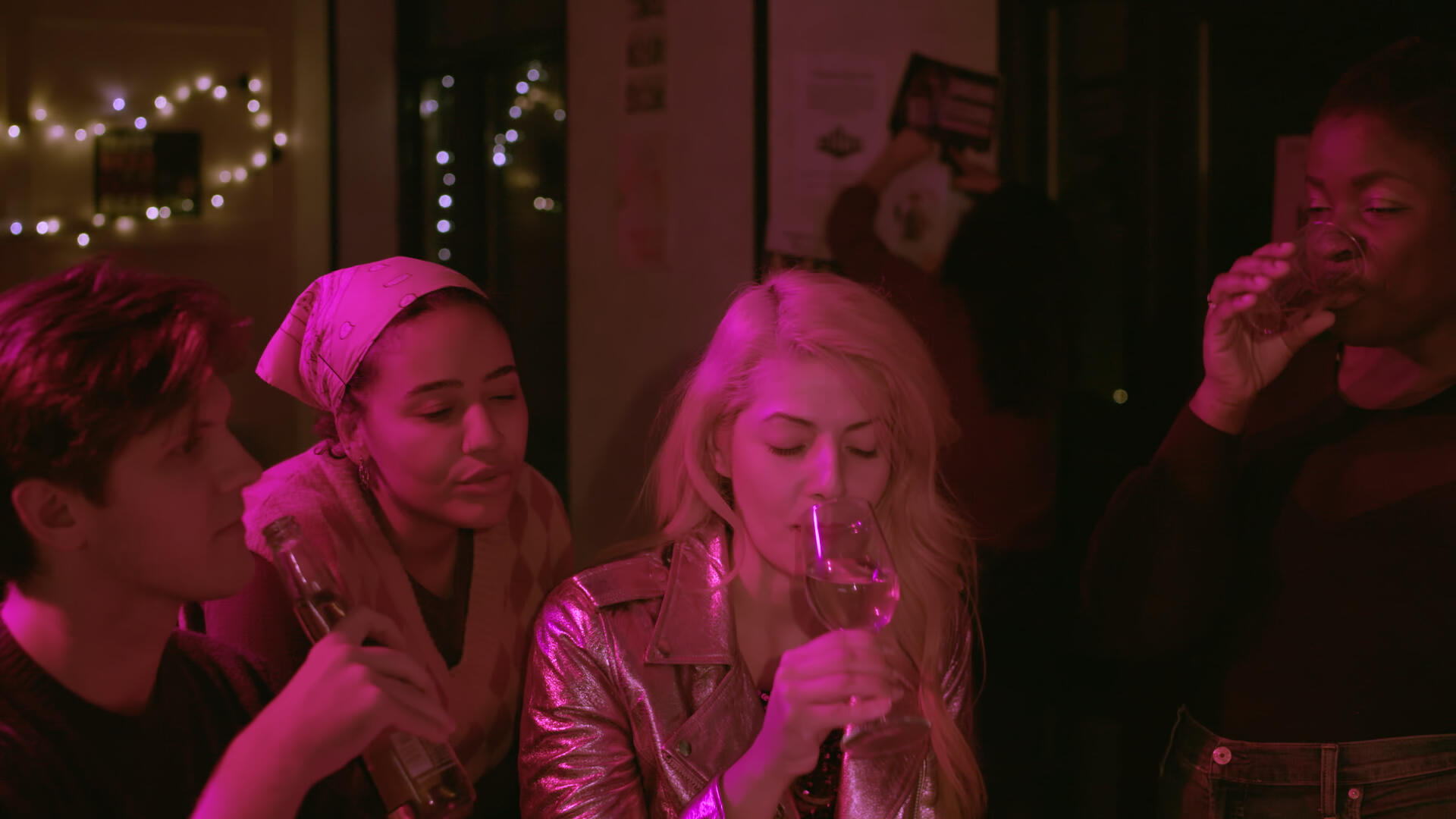 Late Bloomers Still 3 Four leads of late bloomers sit and drink in a bar, bathed in a saturated, magenta light.