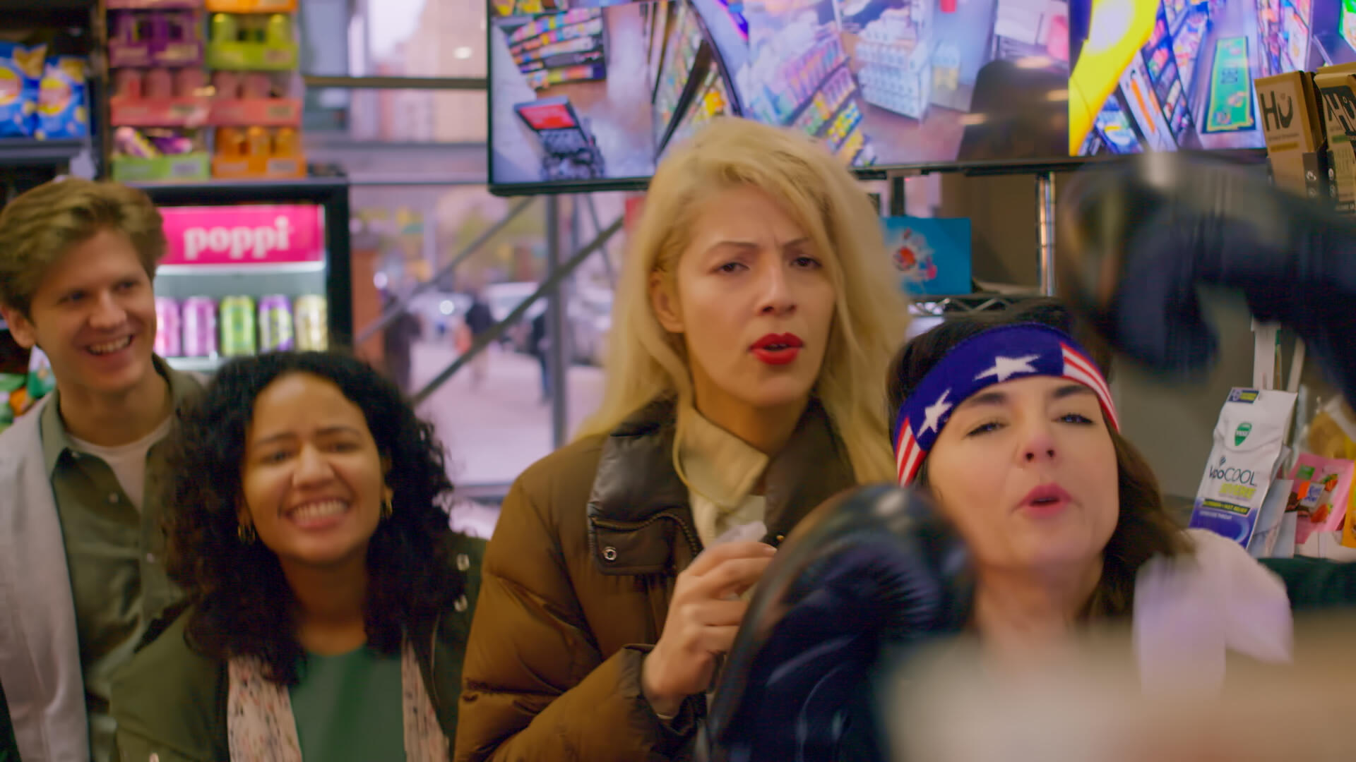 Four of the leads of Late Bloomers in a deli. One woman in front is adorned with boxing gloves and an American flag headband. Three others are behind her in a line, hyping her up.