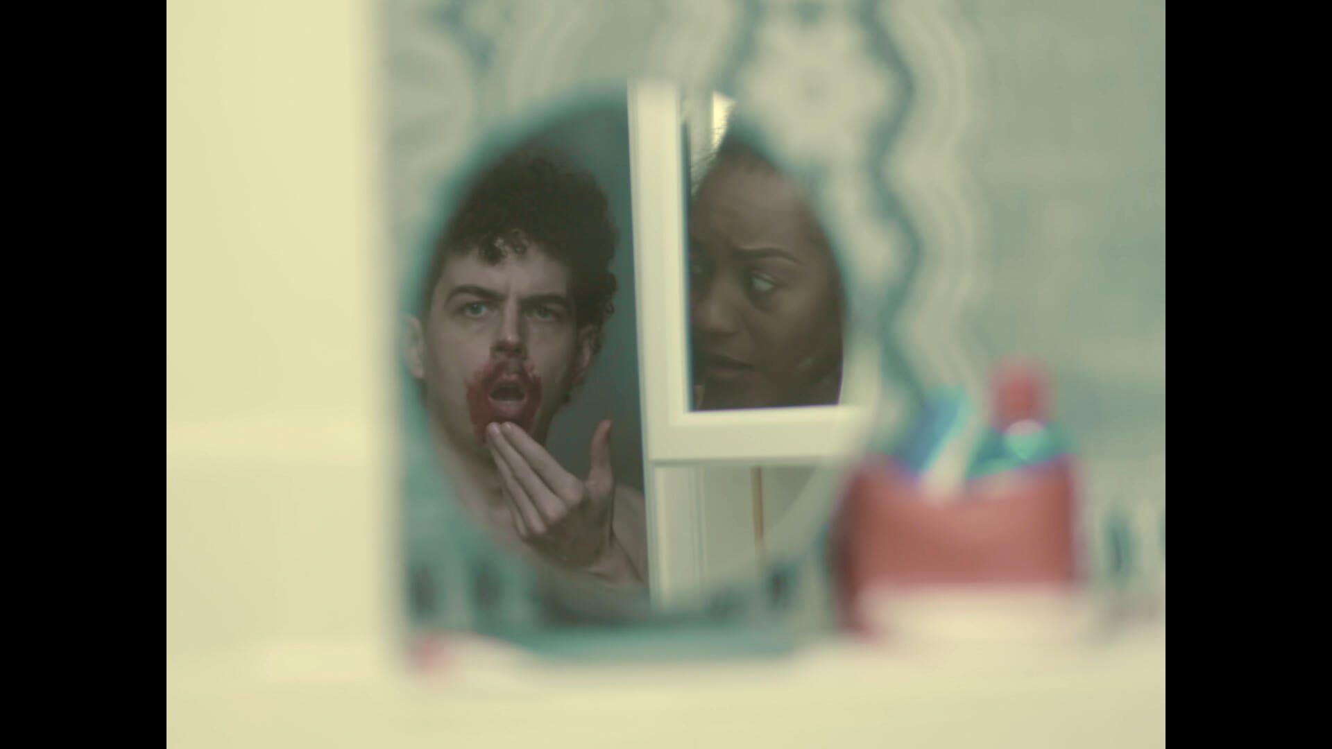 The leading couple stand in the bathroom. The lead actor looks in a mirror on the wall, observing blood around his mouth. The lead actress, reflected in the mirror on the medicine cabinet, looks mortified at her partner.