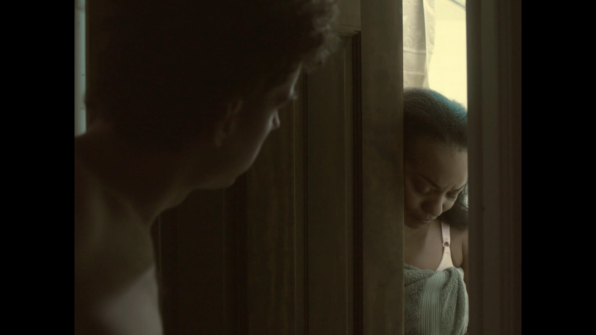 Period. Still 3 The lead actor stands looking at his partner, who is standing behind the bathroom door, looking ashamed at the ground. The scene is lit by the light of the bathroom.