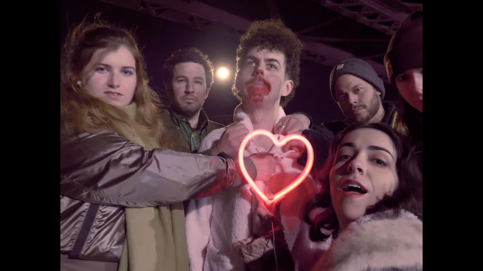 The lead actor stands on the street, blood on his mouth, surrounded by a group of people. One of them holds a neon-lit heart up to him.