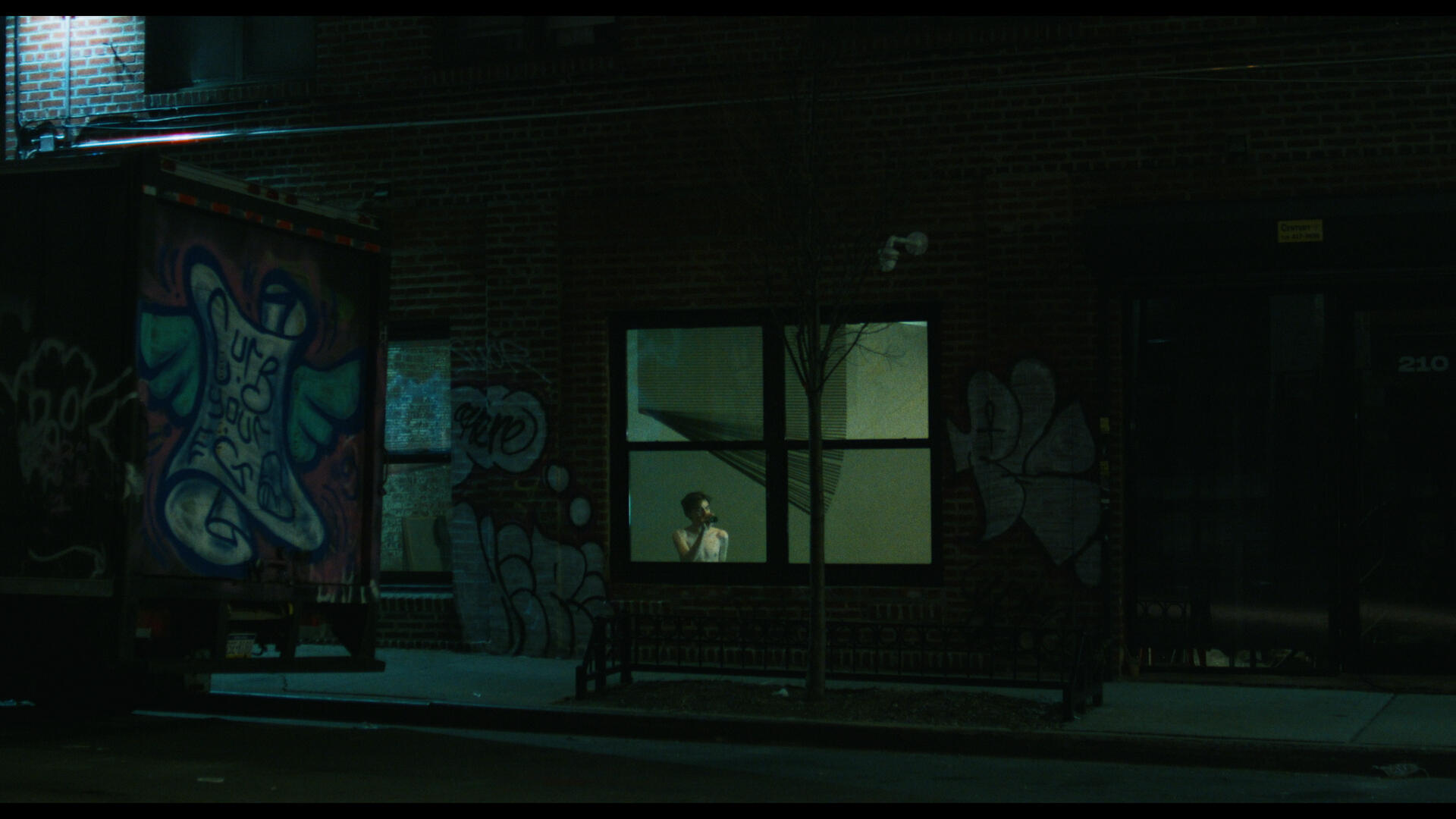 WFTW Still 4 The antagonist of Working for the Weekend sits in a dimly lit room, as seen from across a dark street.